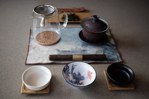 A tea set is arranged on a tabletop, including a transparent glass teapot with a metal lid and handle on a circular wooden mat. There is a lidded ceramic teapot with a red and black pattern, a white bowl, and a small black bowl on wooden coasters. A third bowl is decorated with blue illustrations of fish. Loose tea leaves rest on a small wooden board in the background.