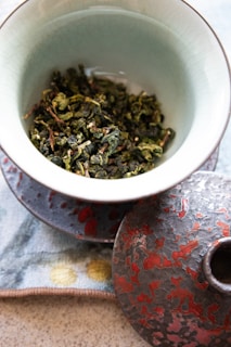 A ceramic bowl filled with dried green leaves, possibly tea leaves, placed on a decorated cloth. The bowl has a matching lid with a textured red and gray design. The setup exudes an artistic and natural aesthetic, with a subtle focus on the textures and colors.