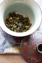 A ceramic bowl filled with dried green leaves, possibly tea leaves, placed on a decorated cloth. The bowl has a matching lid with a textured red and gray design. The setup exudes an artistic and natural aesthetic, with a subtle focus on the textures and colors.