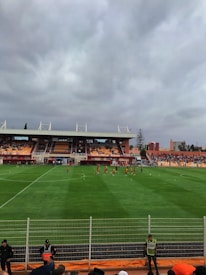 A football stadium with players on the field, spectators in the stands, and cloudy skies overhead. Security personnel are positioned near the fence in front of the stands.
