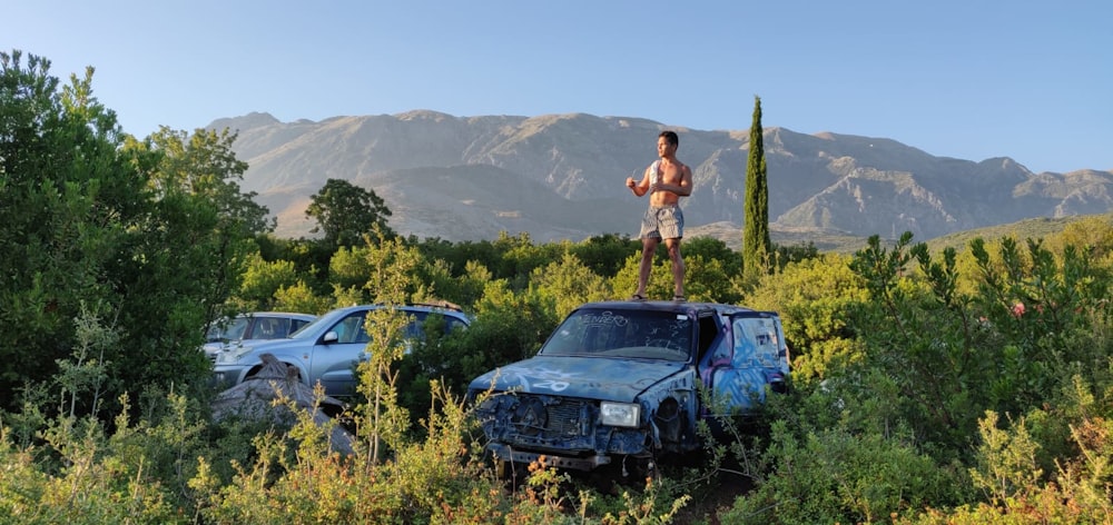 a man standing on a car in a field with trees and mountains in the background