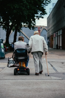 Two elderly individuals are moving along a paved path in an urban setting. One person is using a mobility scooter and the other is walking with a cane. They are both dressed in light-colored clothing and surrounded by buildings and trees.