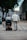 Two elderly individuals are moving along a paved path in an urban setting. One person is using a mobility scooter and the other is walking with a cane. They are both dressed in light-colored clothing and surrounded by buildings and trees.