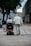 Two elderly individuals are moving along a paved path in an urban setting. One person is using a mobility scooter and the other is walking with a cane. They are both dressed in light-colored clothing and surrounded by buildings and trees.
