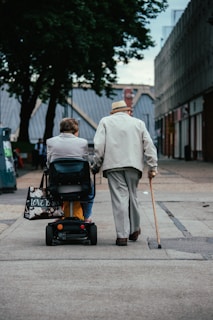 Two elderly individuals are moving along a paved path in an urban setting. One person is using a mobility scooter and the other is walking with a cane. They are both dressed in light-colored clothing and surrounded by buildings and trees.