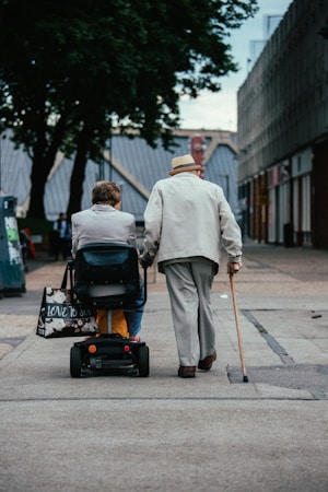 Two elderly individuals are moving along a paved path in an urban setting. One person is using a mobility scooter and the other is walking with a cane. They are both dressed in light-colored clothing and surrounded by buildings and trees.