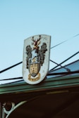 A coat of arms sign featuring a stag with antlers, decorative leaves, and a castle with the word 'WORTH' inscribed at the bottom. The sign is mounted on a structure with a clear blue sky in the background.