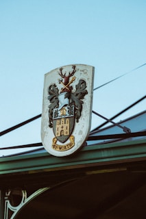 A coat of arms sign featuring a stag with antlers, decorative leaves, and a castle with the word 'WORTH' inscribed at the bottom. The sign is mounted on a structure with a clear blue sky in the background.