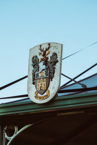 A coat of arms sign featuring a stag with antlers, decorative leaves, and a castle with the word 'WORTH' inscribed at the bottom. The sign is mounted on a structure with a clear blue sky in the background.