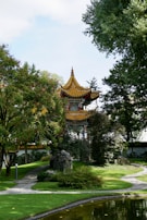 A traditional Asian-style pavilion with a golden roof is nestled among lush green foliage and trees. A winding stone path leads through the well-maintained garden, which borders a serene pond reflecting the landscape.