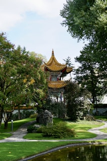 A traditional Asian-style pavilion with a golden roof is nestled among lush green foliage and trees. A winding stone path leads through the well-maintained garden, which borders a serene pond reflecting the landscape.