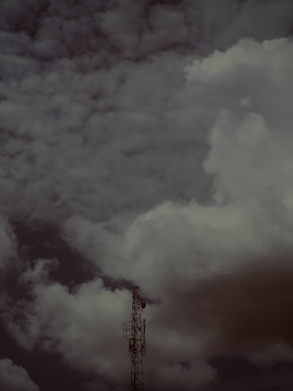 A telecommunications tower stands tall against a backdrop of thick, dark clouds. The sky is overcast, giving a dramatic and moody appearance to the scene.