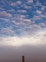 The scene depicts a sky filled with scattered, fluffy clouds against a backdrop of blue. The lower portion shows a gradient transitioning from a brighter horizon to a darker shade toward the bottom. A tall, thin structure rises from the ground, with a set of small lights nearby.