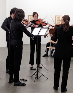 A small string ensemble of four musicians is performing. Each musician is focused on their instrument, with three playing violins and one playing a viola. They are in a minimalist setting likely within a gallery or performance space, with framed artworks on the walls and a few people seated in the background.