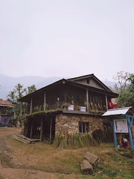 A rustic stone and wooden house stands on a rural dirt path. The structure has an upper and lower level, with grass bundles hanging around the exterior. There are a few small windows, and a corrugated metal roof canopy extends over an area to the side, where a person is partially visible sitting on the ground. The surroundings include patches of grass, trees, and another house visible in the background.