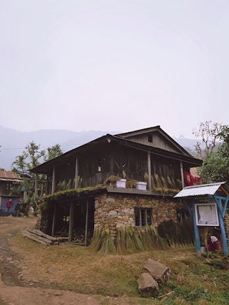 A rustic stone and wooden house stands on a rural dirt path. The structure has an upper and lower level, with grass bundles hanging around the exterior. There are a few small windows, and a corrugated metal roof canopy extends over an area to the side, where a person is partially visible sitting on the ground. The surroundings include patches of grass, trees, and another house visible in the background.