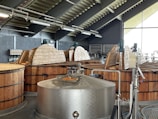 A distillery interior with large wooden fermentation vats and a stainless steel mash tun. The ceiling has exposed beams and industrial lighting. Equipment like pipes and gauges are visible. The room is well-lit by natural light through large windows.