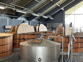 A distillery interior with large wooden fermentation vats and a stainless steel mash tun. The ceiling has exposed beams and industrial lighting. Equipment like pipes and gauges are visible. The room is well-lit by natural light through large windows.