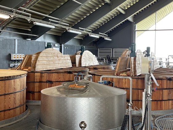 A distillery interior with large wooden fermentation vats and a stainless steel mash tun. The ceiling has exposed beams and industrial lighting. Equipment like pipes and gauges are visible. The room is well-lit by natural light through large windows.