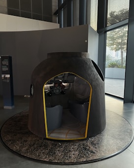 A small, dome-shaped replica or model of a spacecraft capsule with an open entrance and two seats inside. The interior includes control panels and some equipment typically found in spacecraft. The capsule is placed on a platform that resembles a lunar surface. There are large windows and a tall ceiling in the background, indicating the setting is indoors, possibly in a museum or exhibition space.