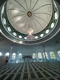 A beautifully designed mosque interior featuring a large, intricately decorated dome with Arabic calligraphy. A central chandelier hangs prominently from the dome. The space below has a green carpet with geometric patterns and is surrounded by arched windows allowing natural light to illuminate the room.