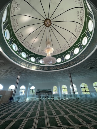 A beautifully designed mosque interior featuring a large, intricately decorated dome with Arabic calligraphy. A central chandelier hangs prominently from the dome. The space below has a green carpet with geometric patterns and is surrounded by arched windows allowing natural light to illuminate the room.