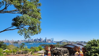 A vibrant cityscape featuring a prominent skyline with skyscrapers and a distinct arch bridge over a large body of water. The scene is framed by lush greenery and trees, with clear blue skies above. People appear to be gathered near a structure with colorful decorations in the foreground.