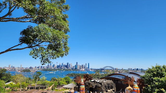 A vibrant cityscape featuring a prominent skyline with skyscrapers and a distinct arch bridge over a large body of water. The scene is framed by lush greenery and trees, with clear blue skies above. People appear to be gathered near a structure with colorful decorations in the foreground.
