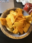 A wooden bowl filled with crispy, golden potato chips. The chips appear seasoned and slightly oily, with a textured surface. In the background, there is a portion of a laptop and a red package with the word 'Ketchup' partially visible.