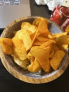 A wooden bowl filled with crispy, golden potato chips. The chips appear seasoned and slightly oily, with a textured surface. In the background, there is a portion of a laptop and a red package with the word 'Ketchup' partially visible.