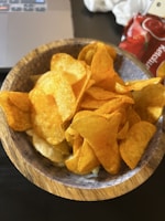 A wooden bowl filled with crispy, golden potato chips. The chips appear seasoned and slightly oily, with a textured surface. In the background, there is a portion of a laptop and a red package with the word 'Ketchup' partially visible.