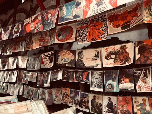 A display of vintage propaganda posters hanging on a wall, featuring various illustrations and slogans. The posters depict historical and political themes, showcasing figures in military and industrial settings, space exploration, and patriotic imagery. Many of the posters have a distinct red and black color scheme, highlighting strong and bold graphical elements.