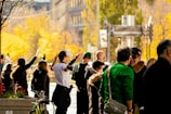 A group of people stand together outdoors in an urban environment with trees and buildings in the background. Some of them are making peace signs with their hands. The scene is brightly lit with warm autumn colors.