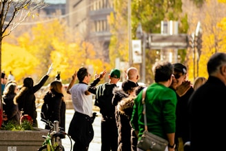 A group of people stand together outdoors in an urban environment with trees and buildings in the background. Some of them are making peace signs with their hands. The scene is brightly lit with warm autumn colors.
