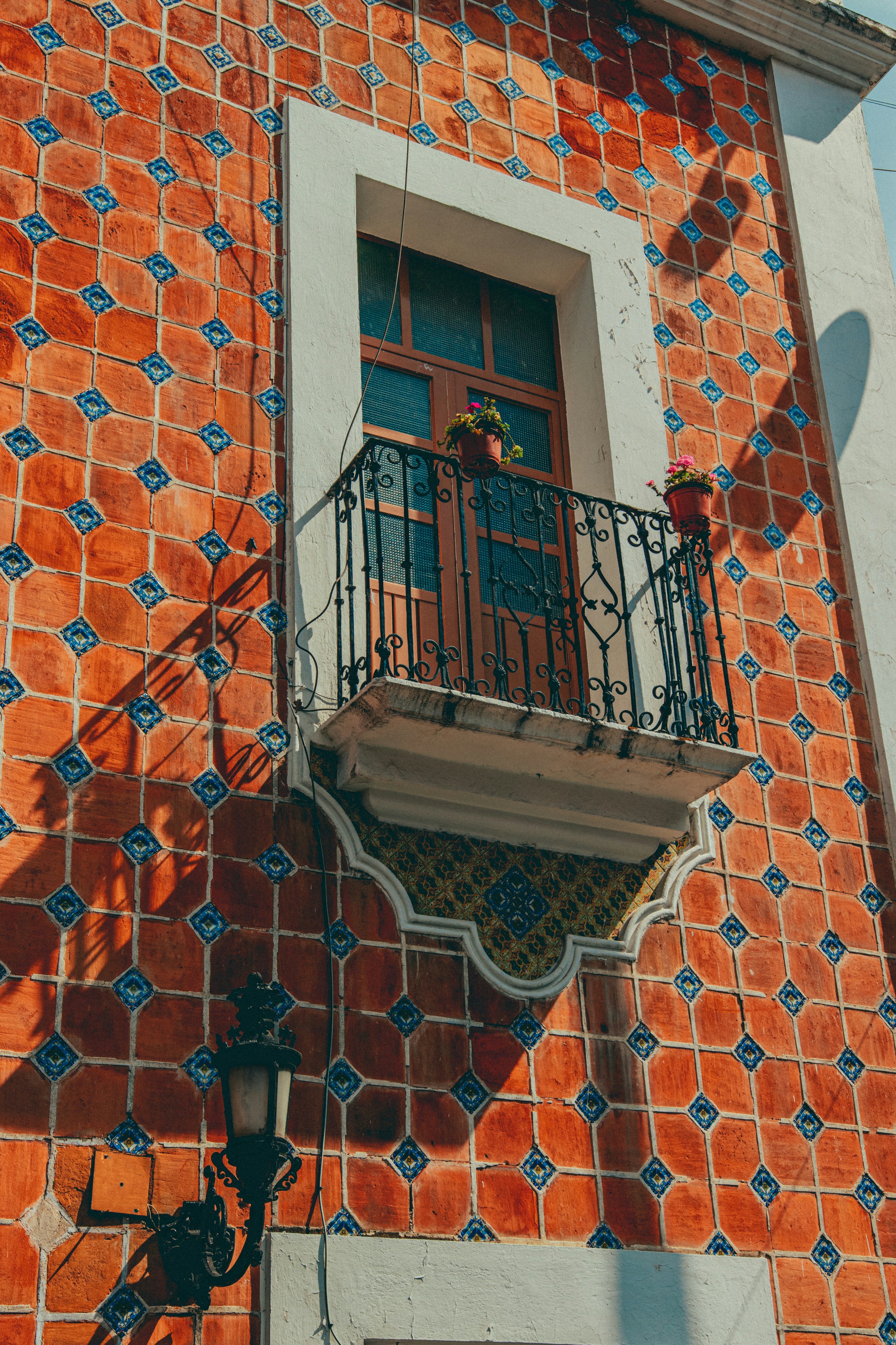 Balcony with wrought-iron railing and two potted flowers sits on a vivid terracotta tile wall, framed by a window and warm sunlight.