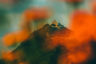 A vibrant hilltop church surrounded by lush greenery with a blurred foreground of orange foliage. The structure is set against a dramatic, overcast sky, creating a contrast between the colorful elements and the dark backdrop.