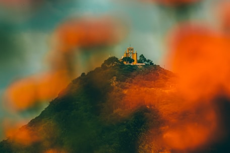 A vibrant hilltop church surrounded by lush greenery with a blurred foreground of orange foliage. The structure is set against a dramatic, overcast sky, creating a contrast between the colorful elements and the dark backdrop.