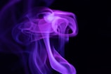 Swirling, ethereal purple smoke against a black background with soft, flowing contours and varying shades of violet.