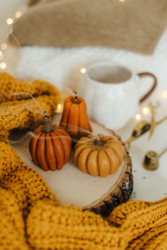 Three small pumpkin-shaped candles are placed on a wooden slice, surrounded by a mustard yellow knitted blanket. In the background, there is a white ceramic mug with a brown handle and various small decorative items. The scene is warmly lit, with small fairy lights adding a cozy ambiance.