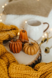 Three small pumpkin-shaped candles are placed on a wooden slice, surrounded by a mustard yellow knitted blanket. In the background, there is a white ceramic mug with a brown handle and various small decorative items. The scene is warmly lit, with small fairy lights adding a cozy ambiance.