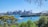 A clear view of a vibrant city skyline with a variety of modern skyscrapers seen across a body of water. Several boats and yachts are sailing on the blue waters. The iconic structure of the Sydney Opera House is visible along with the Sydney Harbour Bridge. Lush green trees frame the foreground of the image, adding a natural contrast to the urban elements.
