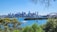 A clear view of a vibrant city skyline with a variety of modern skyscrapers seen across a body of water. Several boats and yachts are sailing on the blue waters. The iconic structure of the Sydney Opera House is visible along with the Sydney Harbour Bridge. Lush green trees frame the foreground of the image, adding a natural contrast to the urban elements.