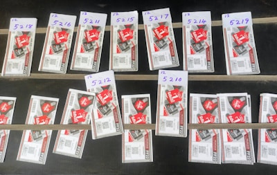 Several rows of paper tickets are arranged on a flat dark surface. Each ticket features a prominent red design with text and barcodes. The tickets are grouped in sets with handwritten labels above them, displaying numbers like '5218', '5216', and others.