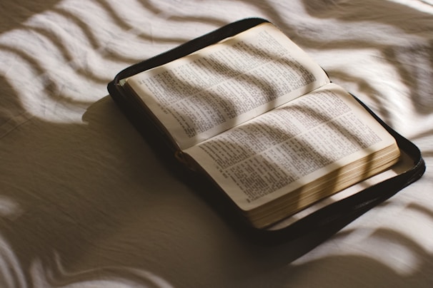 An open book lying on a bed with gentle light and shadow patterns cast across the pages, suggesting a peaceful and contemplative atmosphere.