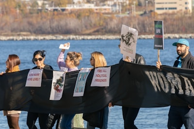 A group of people hold a long black banner with various protest signs, including messages about political prisoners and human rights. The individuals appear to be participating in a peaceful demonstration near a body of water, with buildings visible in the background.