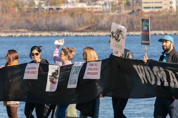 A group of people hold a long black banner with various protest signs, including messages about political prisoners and human rights. The individuals appear to be participating in a peaceful demonstration near a body of water, with buildings visible in the background.
