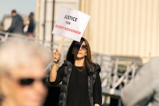 A person wearing sunglasses holds a sign that reads 'Justice for Bloody November' during a protest. The background shows blurry figures and structures, indicating an outdoor environment.