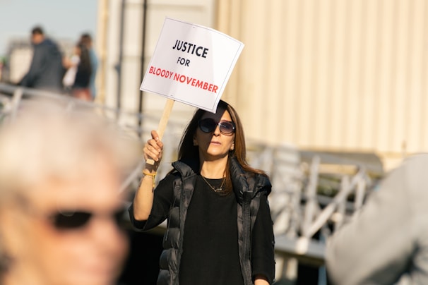 A person wearing sunglasses holds a sign that reads 'Justice for Bloody November' during a protest. The background shows blurry figures and structures, indicating an outdoor environment.