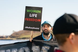 A man holding a sign that reads 'WOMEN LIFE FREEDOM #MahsaAmini'. He is wearing a blue cap and appears to be part of a protest or rally. There is another person partially visible in the foreground, and a body of water with a grassy hill in the background.