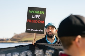 A man holding a sign that reads 'WOMEN LIFE FREEDOM #MahsaAmini'. He is wearing a blue cap and appears to be part of a protest or rally. There is another person partially visible in the foreground, and a body of water with a grassy hill in the background.